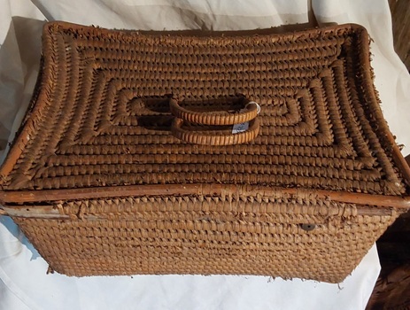 A large lidded firm natural coloured raffia woven basket with two bound plastic woven cane type hinged handles on the lid attached by two pieces of plastic for portability. 