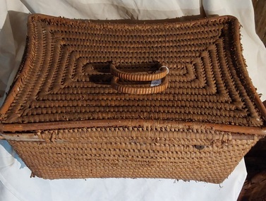 A large lidded firm natural coloured raffia woven basket with two bound plastic woven cane type hinged handles on the lid attached by two pieces of plastic for portability. 