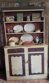 A tall wooden handmade cream and brown kitchen dresser with two framed shelves built on top of the bench, two brown drawers with round wooden knobs and two opening doors.
