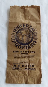 A vintage tall narrow rectangular brown paper bag with side gussets for greater storage. The large brand symbol Universal Providers for the N.H. Hobbs store in Bemboka is printed on the front.