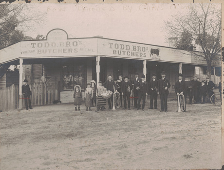 Black and white image of members of a brass band standing outside of a butcher shop.