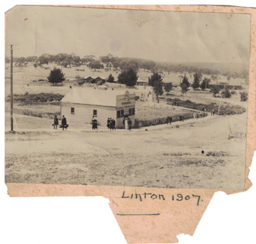 Photograph of a wooden building with railway station behind, passengers from train walking up street toward township. 