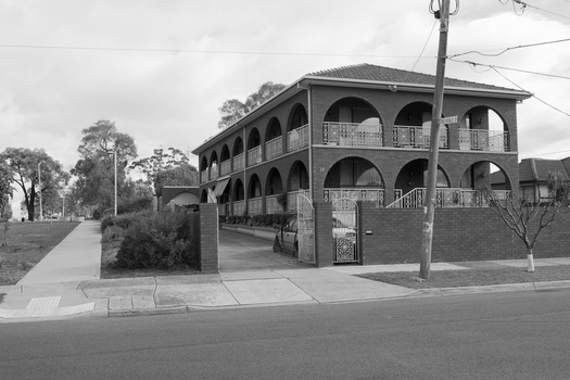 Black and white photograph of a street corner, with a two storey brick house. The house has a series of wide brick arches on both sides and floors, with wire lattice fencing between the arches.