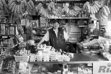 Black and white photograph of a man standing behind the counter of an Asian grocery store. The counter has piles of plastic containers with food products, a stand of confectionery to the left and bunches of bananas hang above the counter top. 