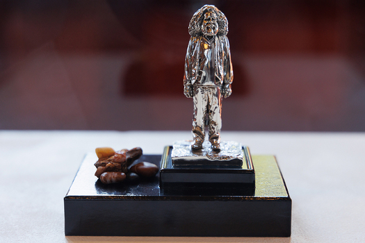 A small silver sculpture of a person with bushy hair and a beard dressed in baggy clothing. The figure stands on a dark plinth with a selection of small rocks to the left of the figure. 