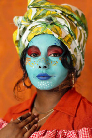 A portrait of a woman with her face painted in bright colours. Her lips and eyes have dramatic outlines; she is wearing a patterned headscarf and looks into the camera with one hand resting on her collar bone. 