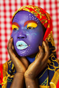 A portrait of a woman whose face is painted in bright colours. She wears a colourful, patterned scarf on her head, and the backdrop is a checkered fabric in soft focus. Her hands are held up near her face and her gaze is directed to the side of the frame. 