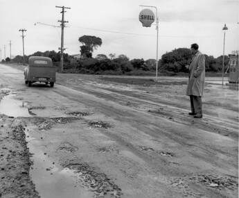 A man, dressed in a trench coat and holding a pipe, watching as a car drives past along the muddy, potholed road
