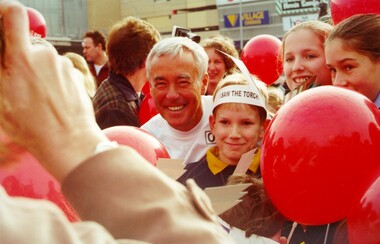 A smiling man, dressed in white, surrounding by smiling children holding red balloons