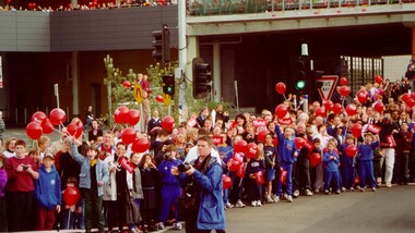 A crowd of mostly children, holding red balloons, wait for the Olympic torch relay to pass by