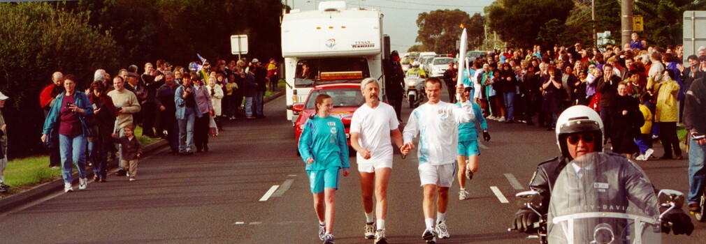 Olympic torch bearers run along the road with a crowd on either side cheering them on