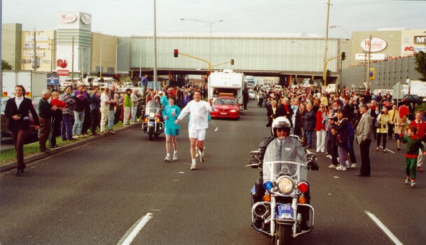 Olympic torch bearer and support team run along the road carrying the torch and surrounded by spectators