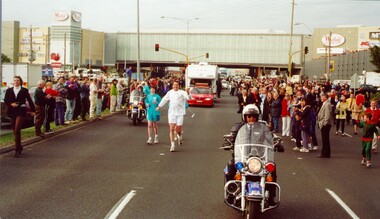 Olympic torch bearer and support team run along the road carrying the torch and surrounded by spectators