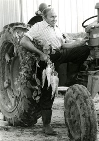 A man standing next to a tractor with one foot on the ground and the other on the tractor step, holding a bunch of parsnips