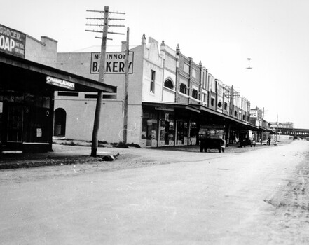 A long view down McKinnon Road taking in a shopping strip and ending with a passenger train going through the level crossing while the gates are closed