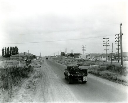 A truck driving along a sealed road while a car waits on a side road 