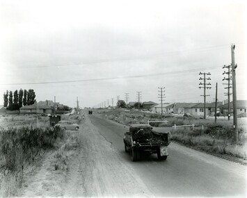 A truck driving along a sealed road while a car waits on a side road 