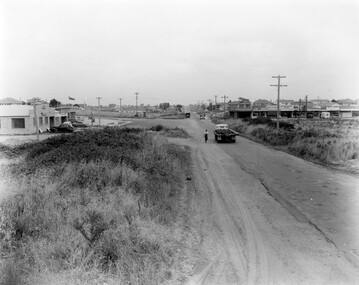 A sealed road in poor condition runs through Moorabbin towards a shopping precinct