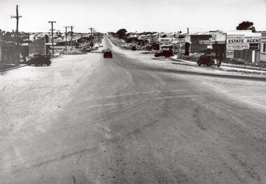 A poorly sealed road with wide gravel shoulders runs through the shopping strip of Moorabbin
