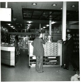 A woman and man standing in front of catalogue drawers inside a library with a second woman behind a desk in the background 