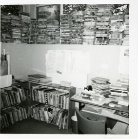 A view of a library corner, with picture books on two low shelves, a desk, and stacks of books above a dividing wall.