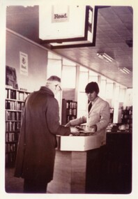 Two men face one another across a library circulation desk. An illuminated sign saying 'Read' hangs above them.