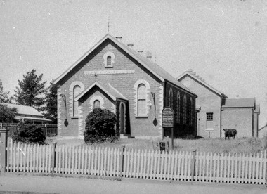 A small church building behind a picket fence. A cow stands to the right of the church in the grass. 