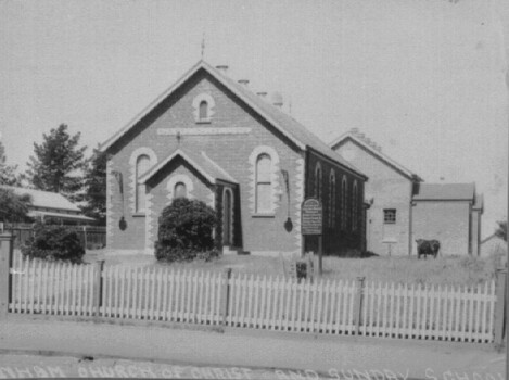 A small church building behind a picket fence. A cow stands to the right of the church in the grass. 