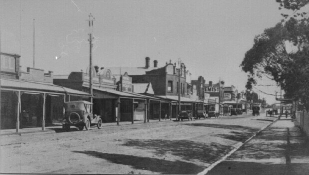 A view of a shopping strip with cars parked along the left hand side of the road. A horse drawn cart can be seen approaching