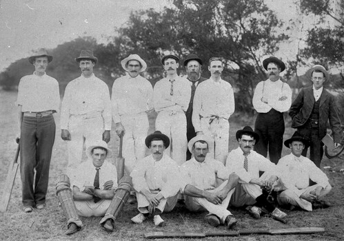 An image of 13 men in a field facing the camera. Most a wearing cricket whites and there are several cricket bats in the image