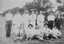 An image of 13 men in a field facing the camera. Most a wearing cricket whites and there are several cricket bats in the image