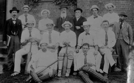 A photograph of 15 men facing the camera in front of a brick wall. 11 are dressed in cricket whites and four in formal attire