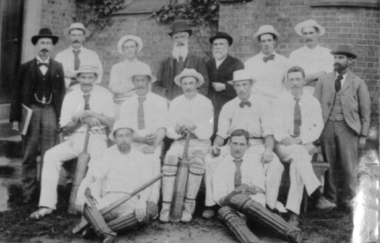 A photograph of 15 men facing the camera in front of a brick wall. 11 are dressed in cricket whites and four in formal attire