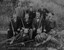 A photograph of nine men facing the camera in an outdoor setting. Six men are holding rifles. 