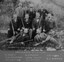 A photograph of nine men facing the camera in an outdoor setting. Six men are holding rifles. 