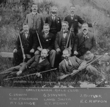 A photograph of nine men facing the camera in an outdoor setting. Six men are holding rifles. 