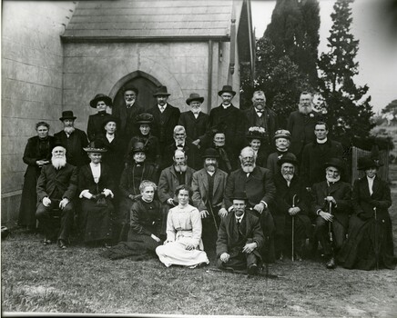 A black and white photograph of a large group of men and women all looking at the camera, in front of a building.