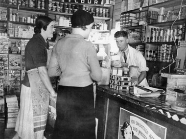 Photograph of a grocery shop interior, with a male behind the counter facing the camera and two females facing the male