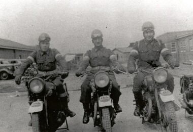 Three men in uniform sit astride three motorcycles. Cars and buildings are in the background of the image