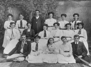 The women cricketers are all dressed in white. Two men are sitting on floor holding crickets bats, a woman dressed in dark colours is sitting among the team, and another man is standing in the back row.