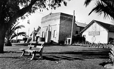 Three buildings abut a grassed area, with trees and a bench seat in front