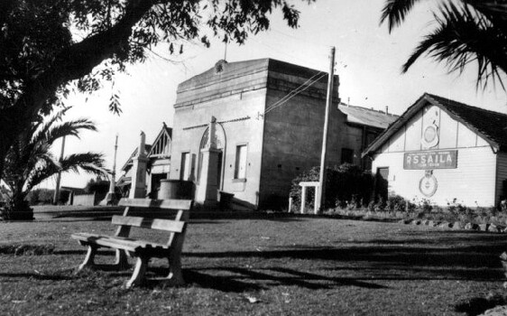Three buildings abut a grassed area, with trees and a bench seat in front