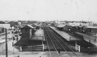 Black and white photograph of a railway line, two platforms and station buildings. Shops and people can be seen behind the station buildings. 