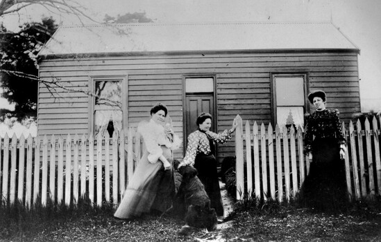 Three women standing outside a small wooden cottage.