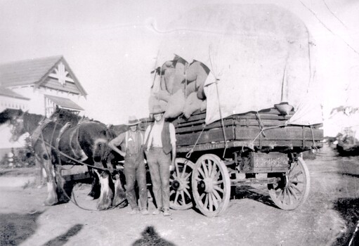 Two men standing in front of a heavily loaded horse-drawn wagon.