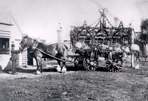 A man in a bowler hat is holding the reins of a horse that is attached to a decorated wagon.