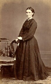 Woman standing beside a chair, wearing a full length dress.