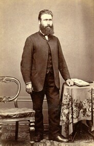 Norman McSwain standing between a chair and a table, dressed in a three piece suit.