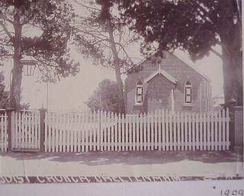 The brick church is set behind a white picket fence, with a gate, and surrounded by large trees.