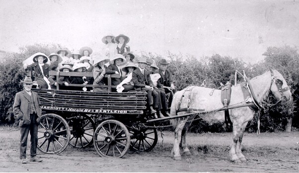 A group of women an dchildren seated in the tray of a horse drawn wagon with three men sitting on the front bench, one holding the reins. There is also a man standing on the ground beside the carriage.